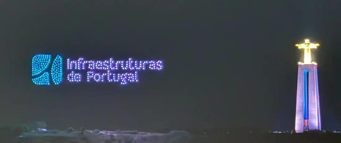 Logótipo da Infraestruturas de Portugal desenhado por drones no céu noturno, ao lado do Cristo Rei iluminado