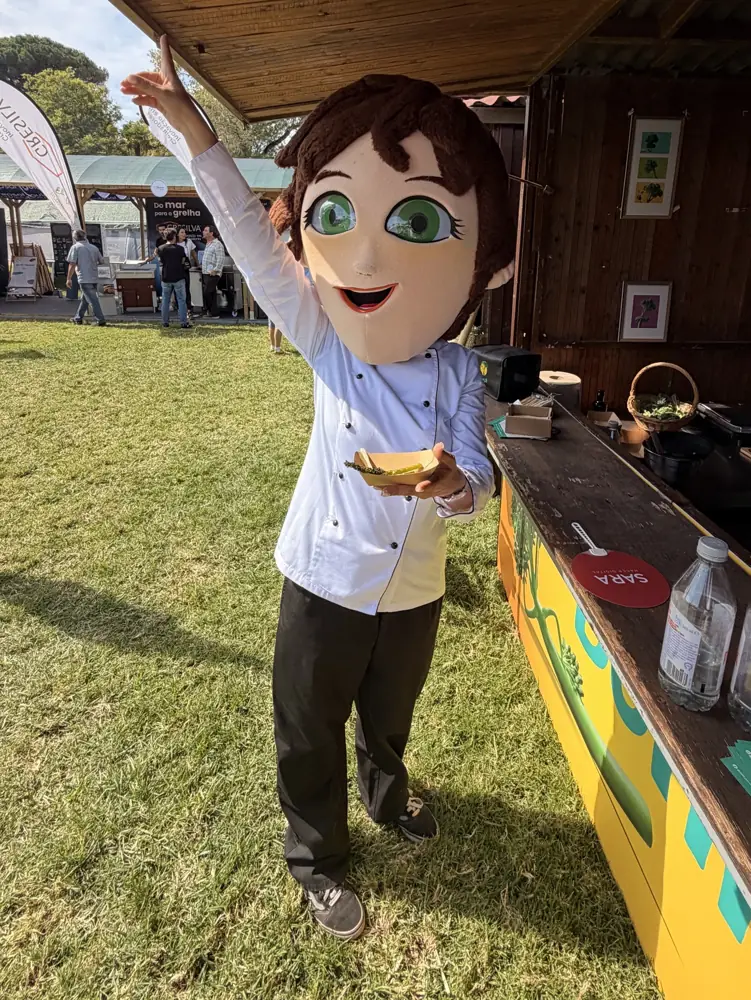 Mascote de chef a acenar junto a um stand numa feira gastronómica ao ar livre