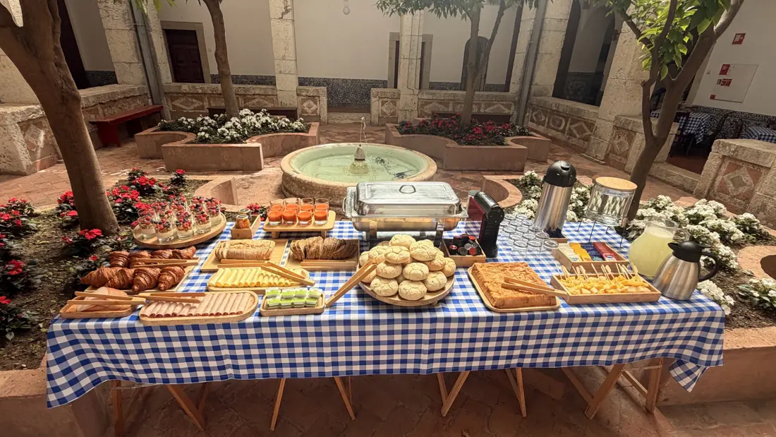 Mesa de pequeno-almoço com toalha aos quadrados azuis num claustro com fonte e flores