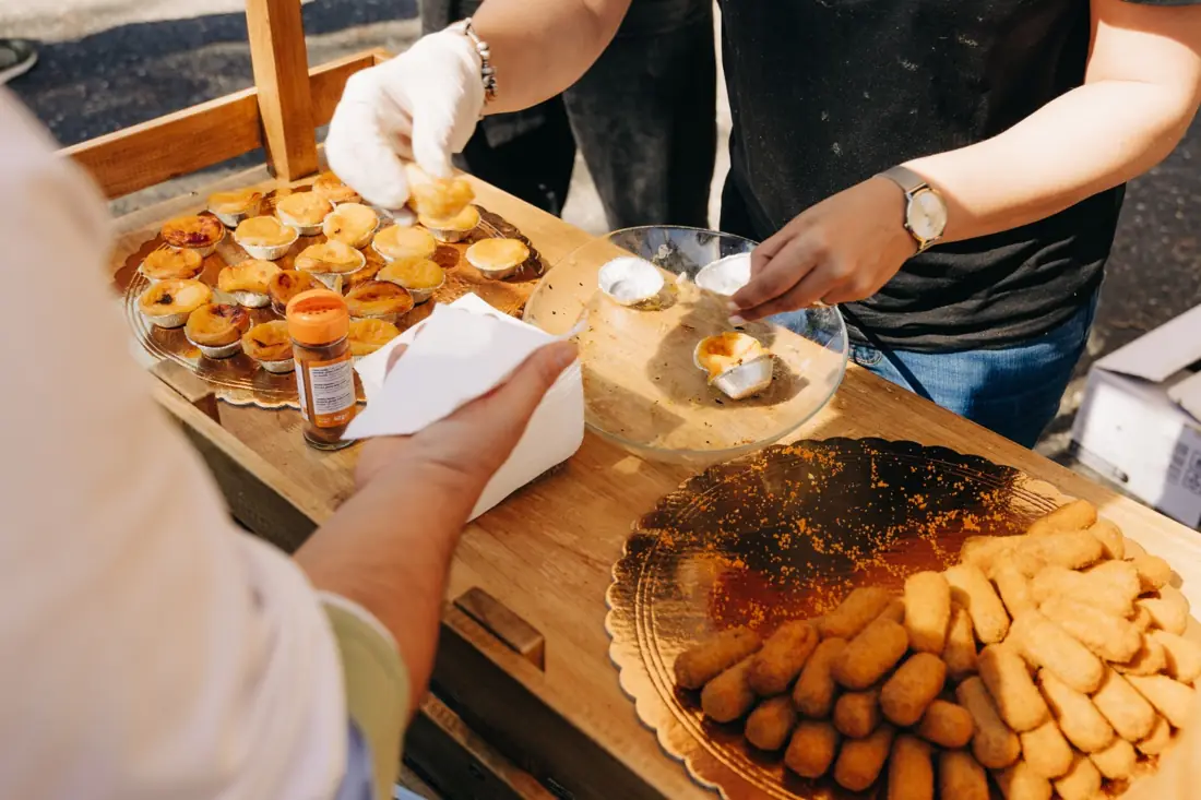 Mãos com luvas a servir pastéis de nata e croquetes num balcão de madeira